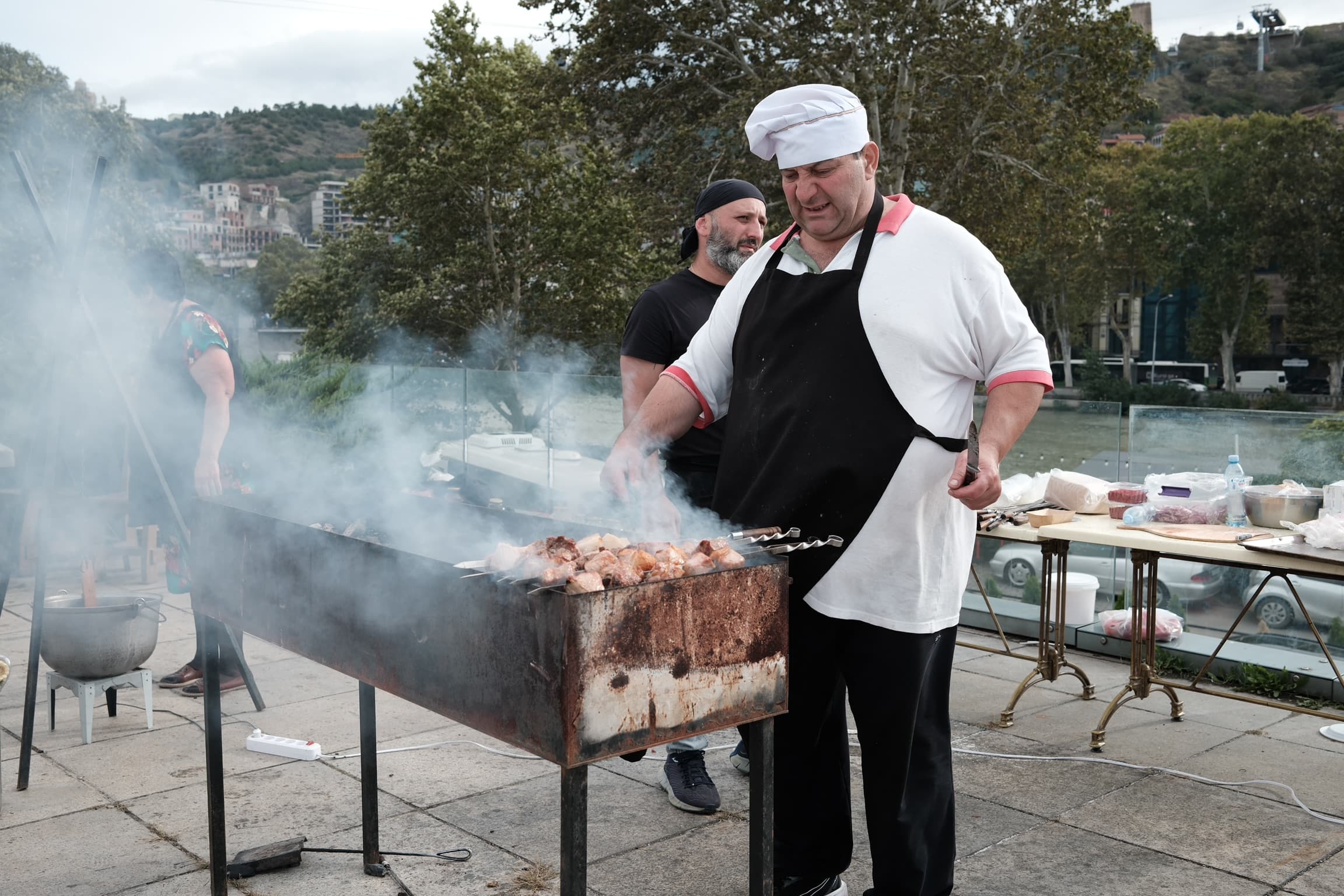 Mtsvadi cook working the grill above the Narikala river - Tbilisi in the background
